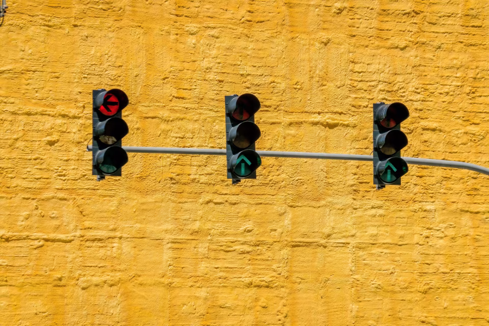 Drei Ampeln vor gelber Wand, links roter Pfeil nach links, mittig und rechts grüne Pfeile nach oben