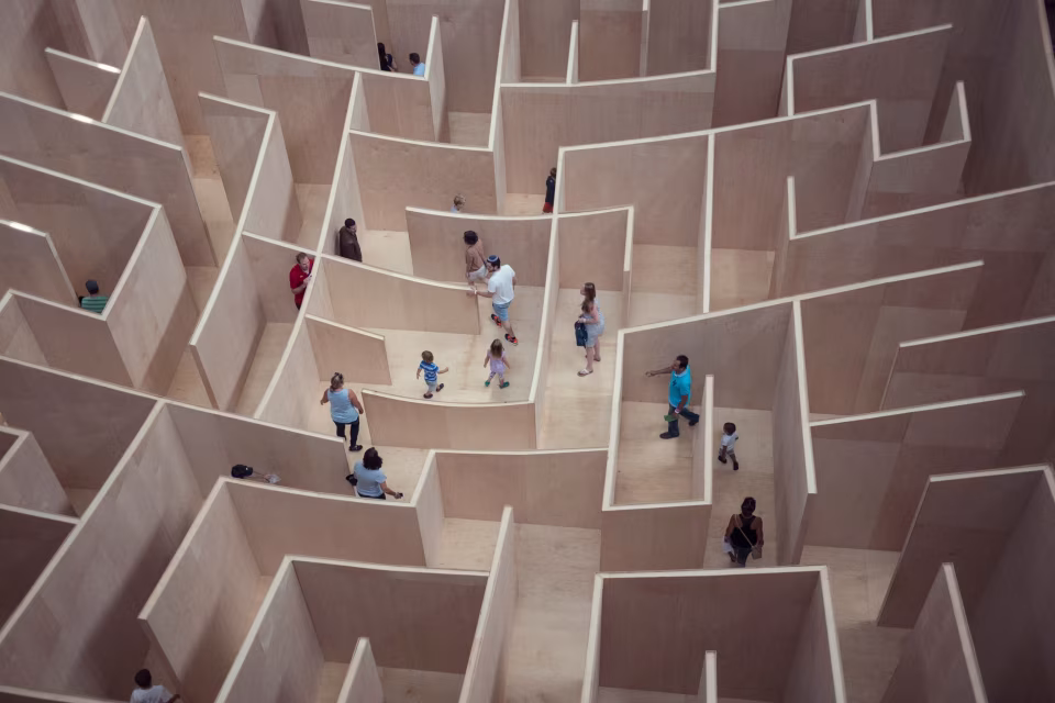 Menschen bewegen sich in einem großen Labyrinth aus hellen Holzplatten mit hohen Wänden, Luftaufnahme von oben